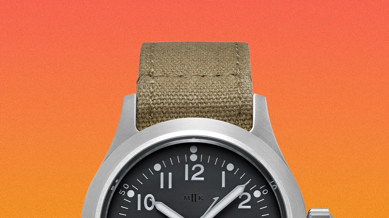 Silver metal wristwatch with black dial, white numerals, and olive green fabric strap on an orange gradient background.