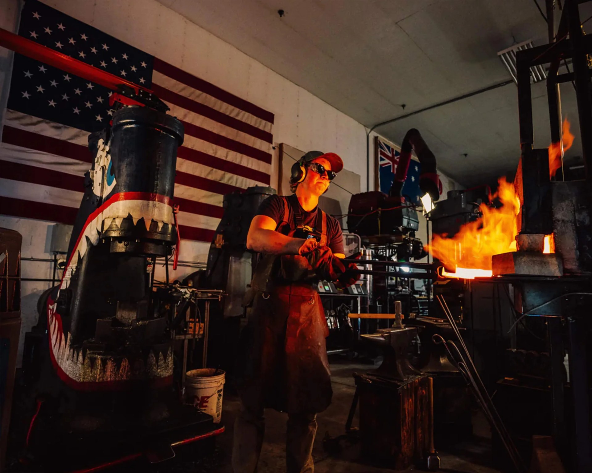 Blacksmith wearing protective gloves, apron, and ear protection working with glowing hot metal in a workshop with American and Australian flags.