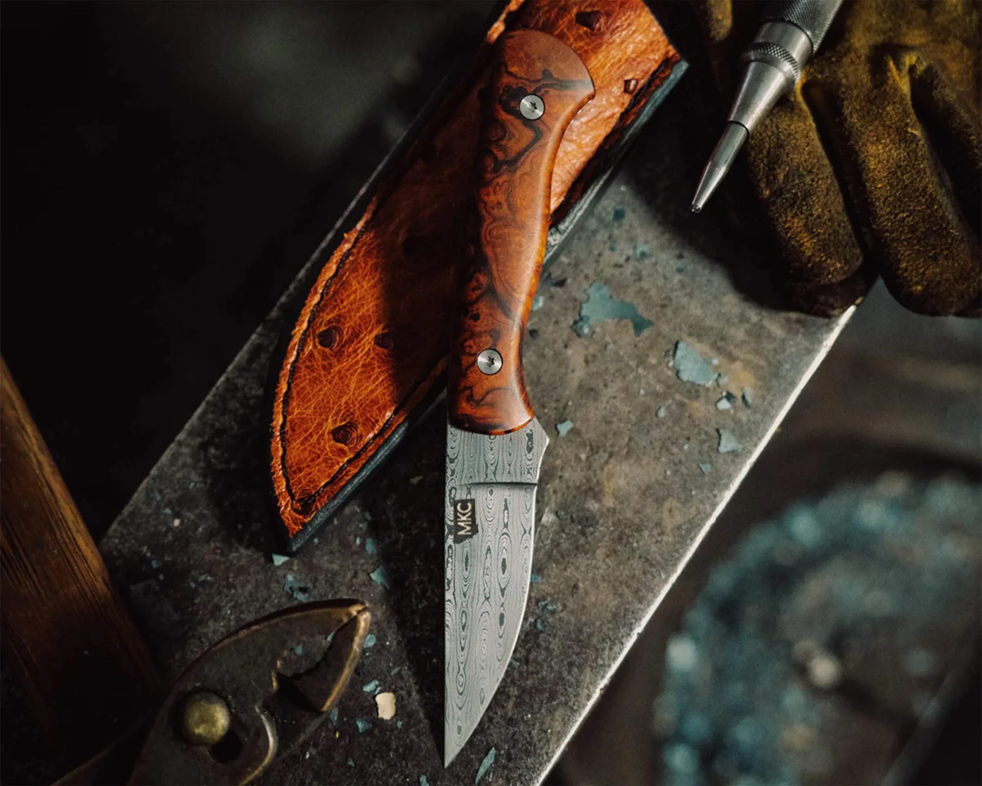 Damascus steel knife with a wooden handle and leather sheath on a metal surface next to a tool and a gloved hand.