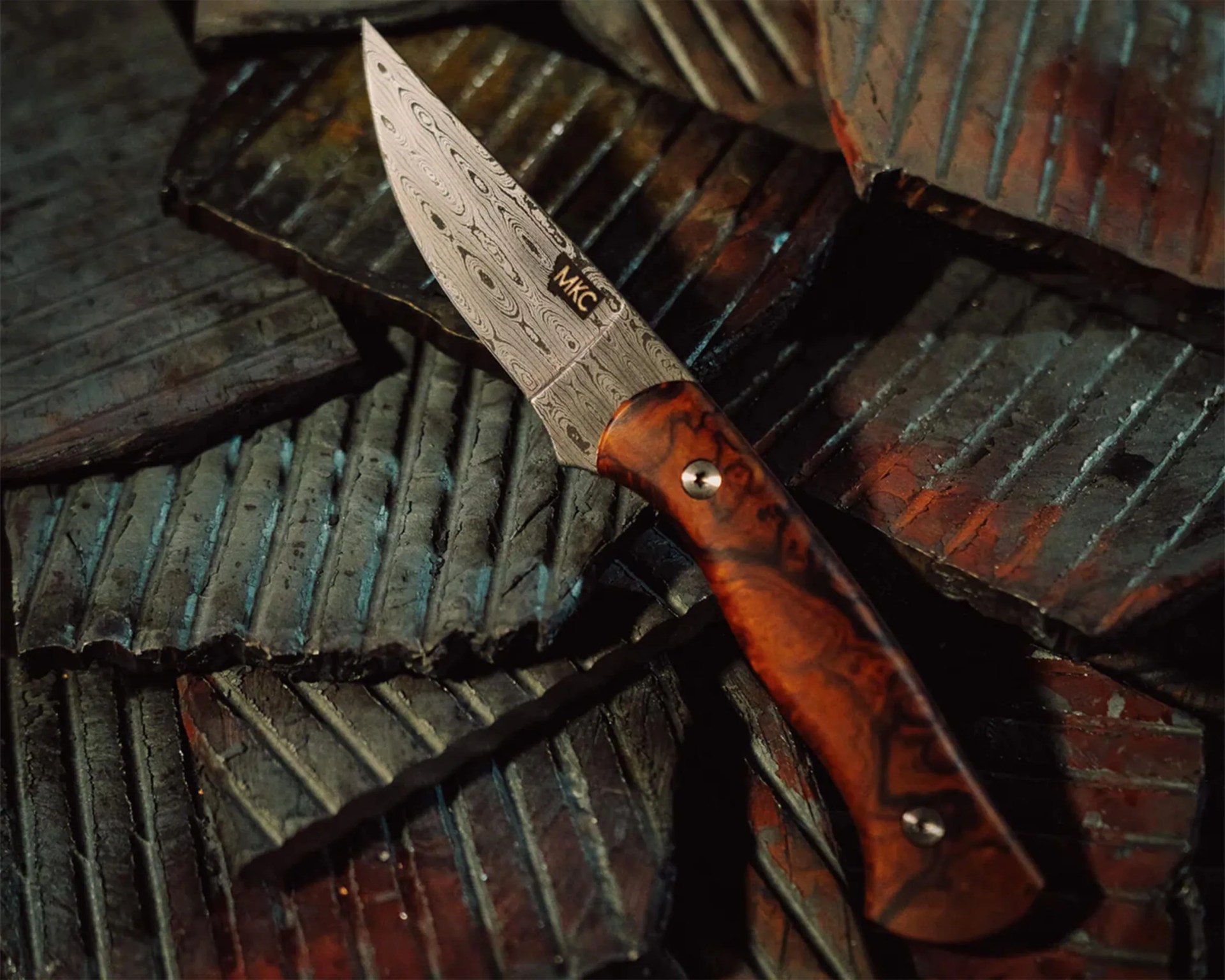 Knife with a patterned Damascus steel blade and a polished wood handle resting on textured metal pieces.