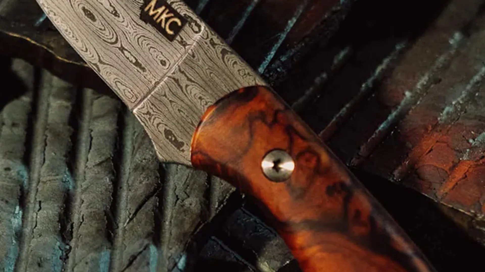 Close-up of a knife with a patterned steel blade marked "MKC" and a polished reddish-brown wooden handle with a metal rivet.