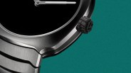 Close-up of a stainless steel watch case with a textured black crown against a teal background.