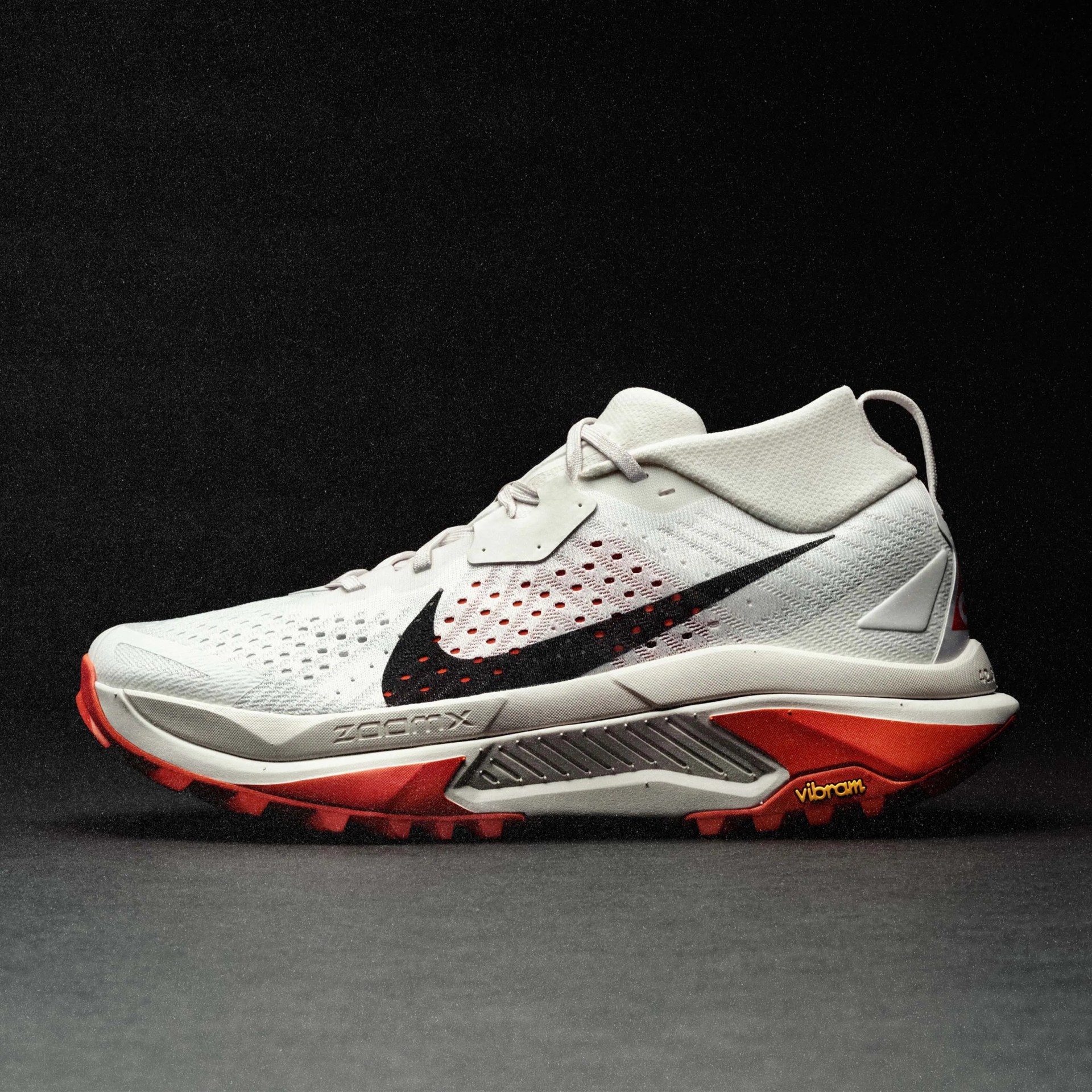White and red Nike ZoomX running shoe with black swoosh and Vibram sole on a dark surface.