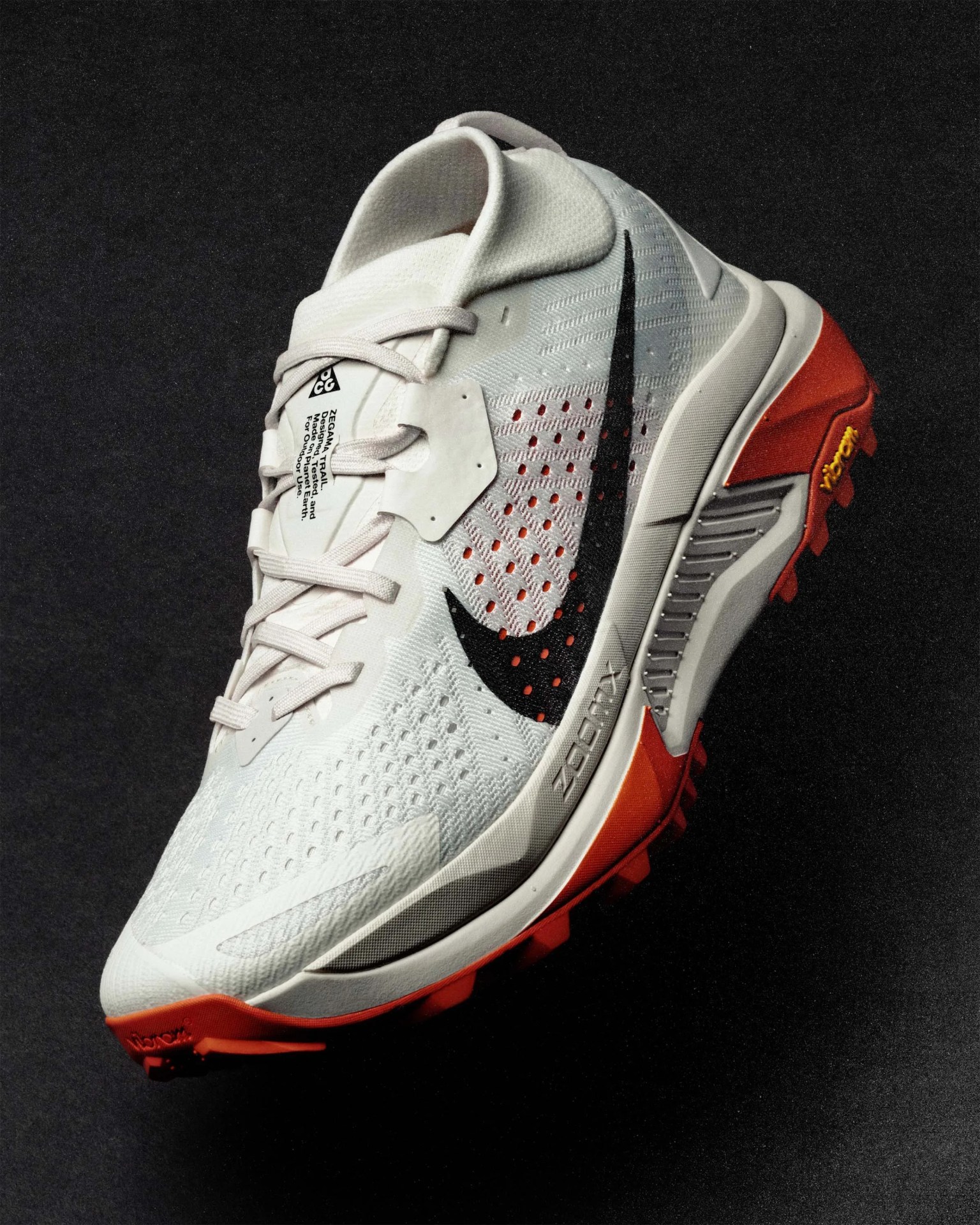 White and orange Nike ACG trail running shoe with black swoosh and Vibram sole on a black background.