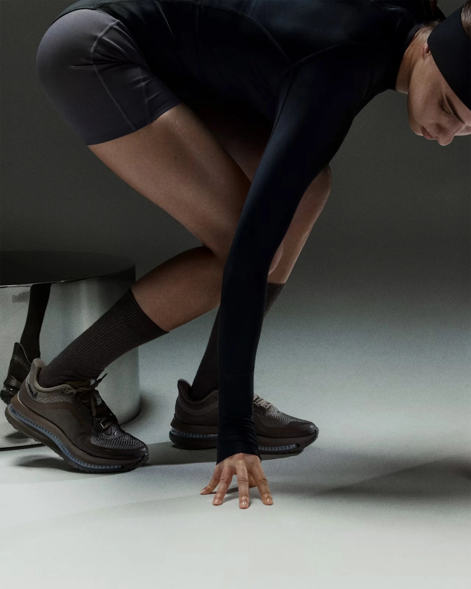Person wearing dark brown athletic shoes, black shorts, and a black long-sleeve top, crouching with one hand on the floor.