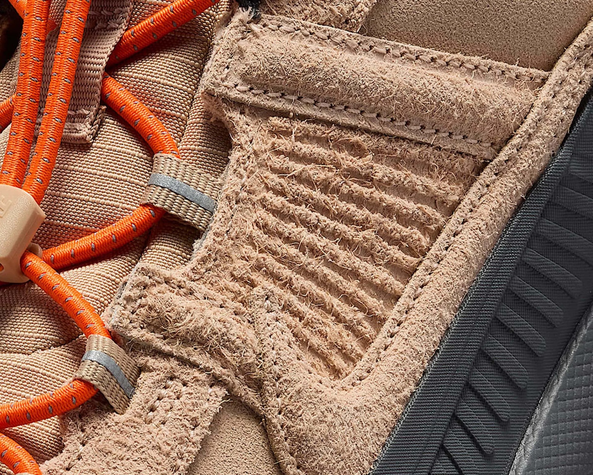 Close-up of tan suede sneaker with textured stitching, orange laces with reflective details, and black rubber sole edge.