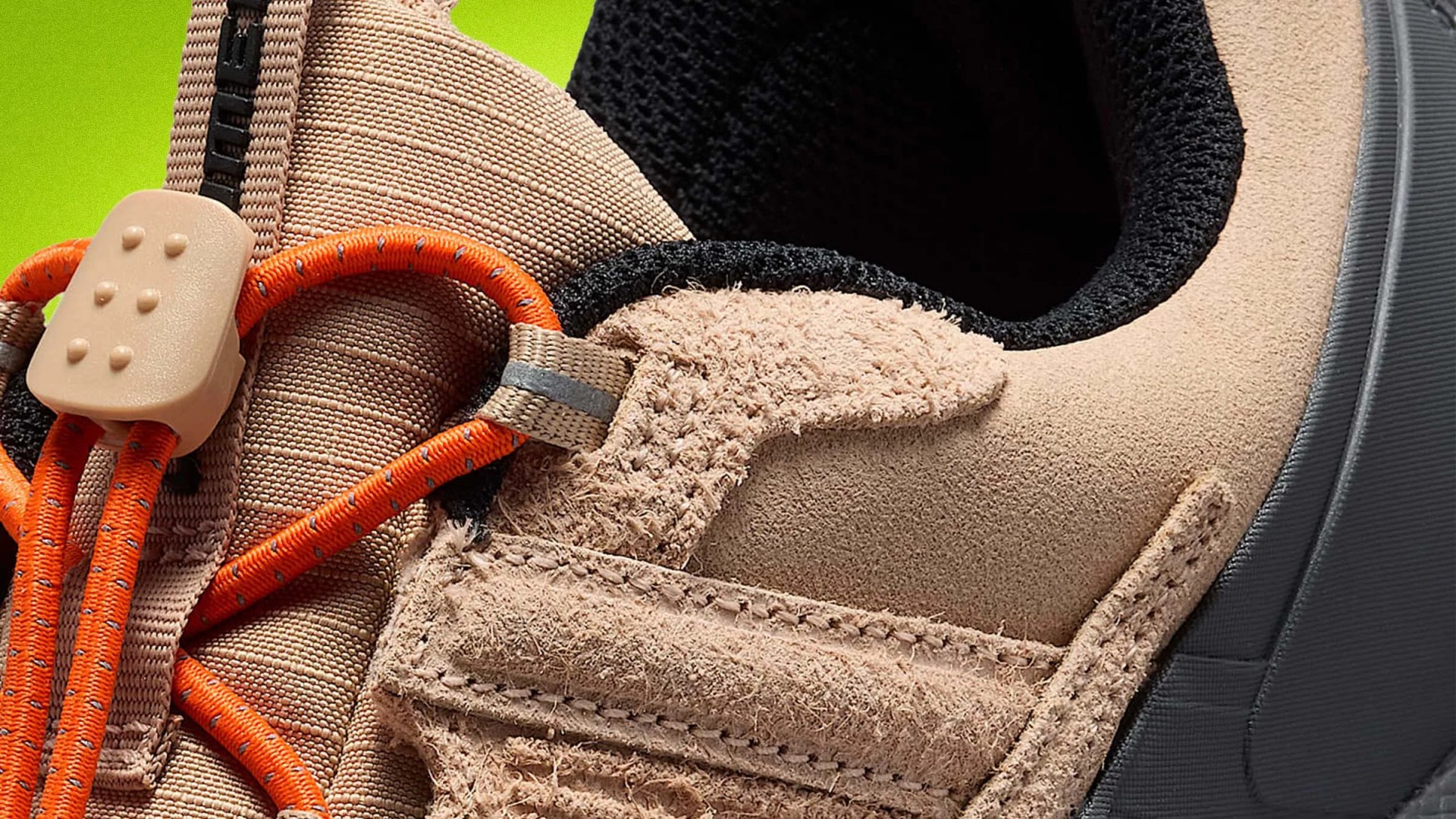 Close-up of tan suede and fabric sneaker with orange laces and black inner lining against a green background.