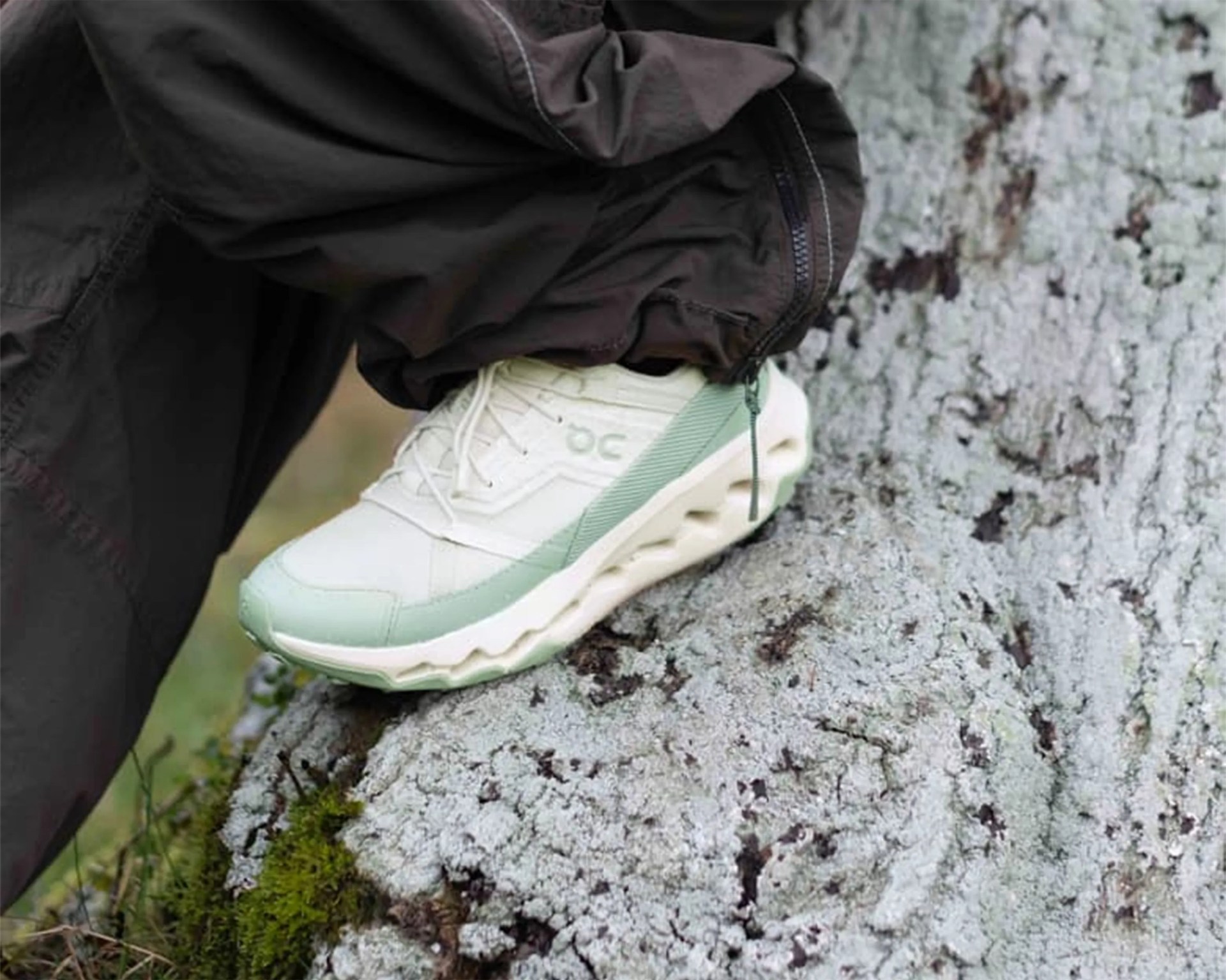 White and light green athletic shoe with textured sole worn with black pants on a mossy rock.