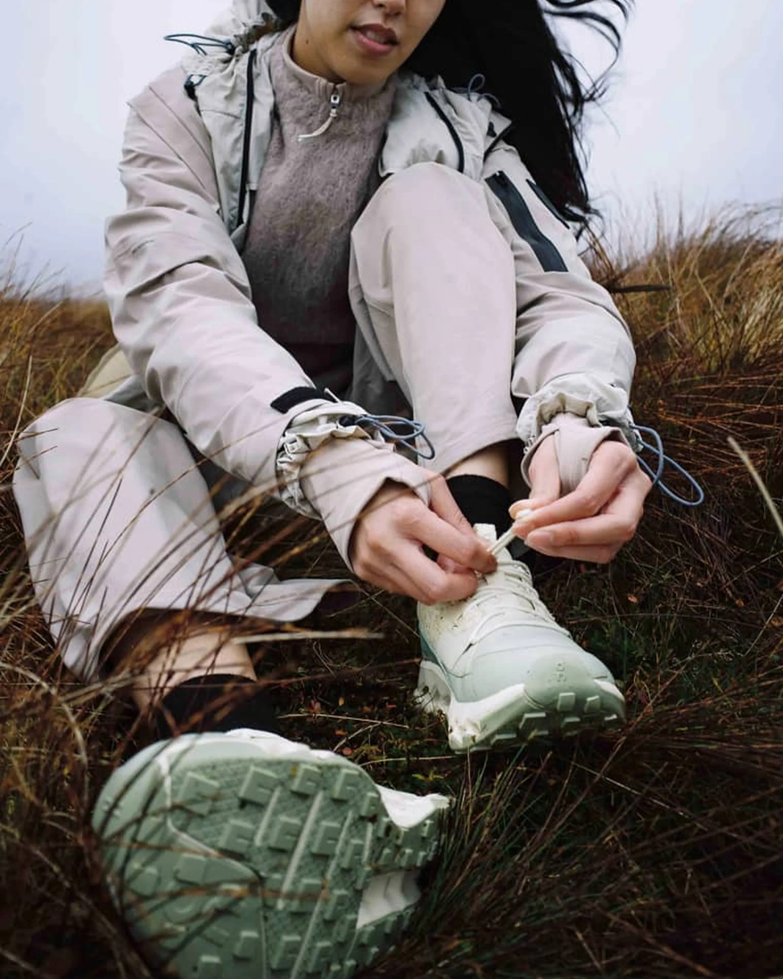 Person wearing light beige outdoor jacket and pants tying white hiking shoes while sitting on grassy terrain.