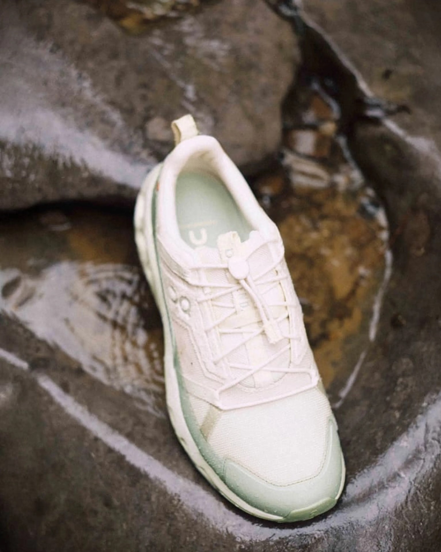 White and light green athletic sneaker with laces, placed on wet rocks.