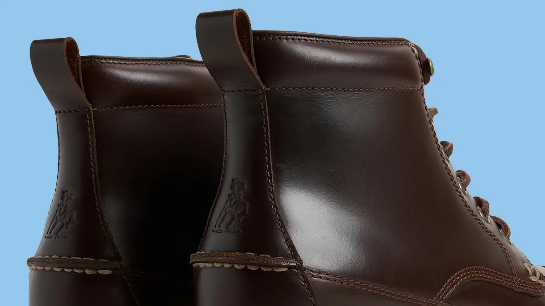 Pair of dark brown leather boots with pull tabs and embossed logo on the heel against a blue background.