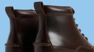 Pair of dark brown leather boots with pull tabs and embossed logo on the heel against a blue background.