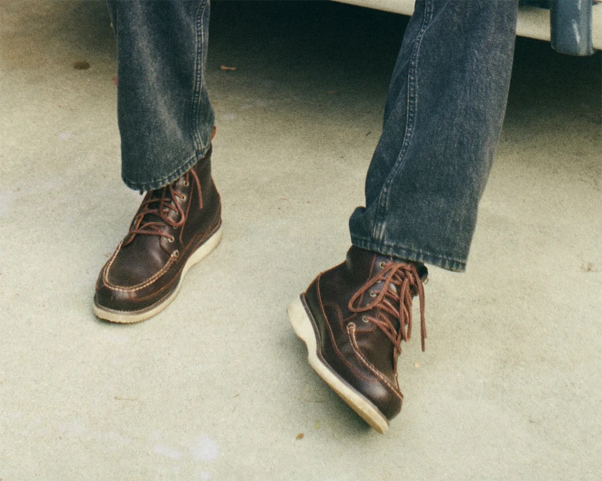 Brown leather moc toe boots with white soles worn with dark blue jeans on concrete floor.
