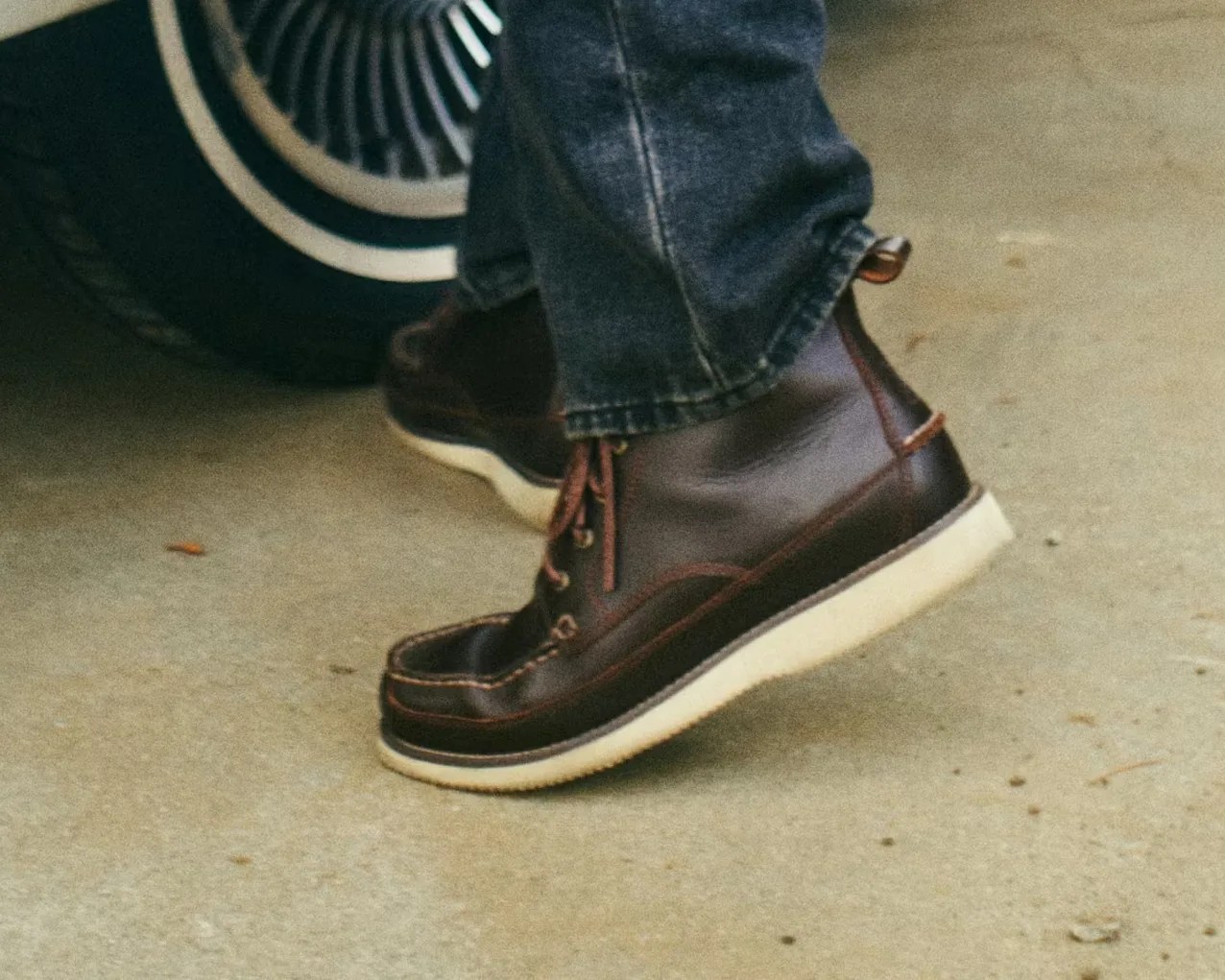 Dark brown leather moc toe boots with white soles worn with dark denim jeans near a car tire.