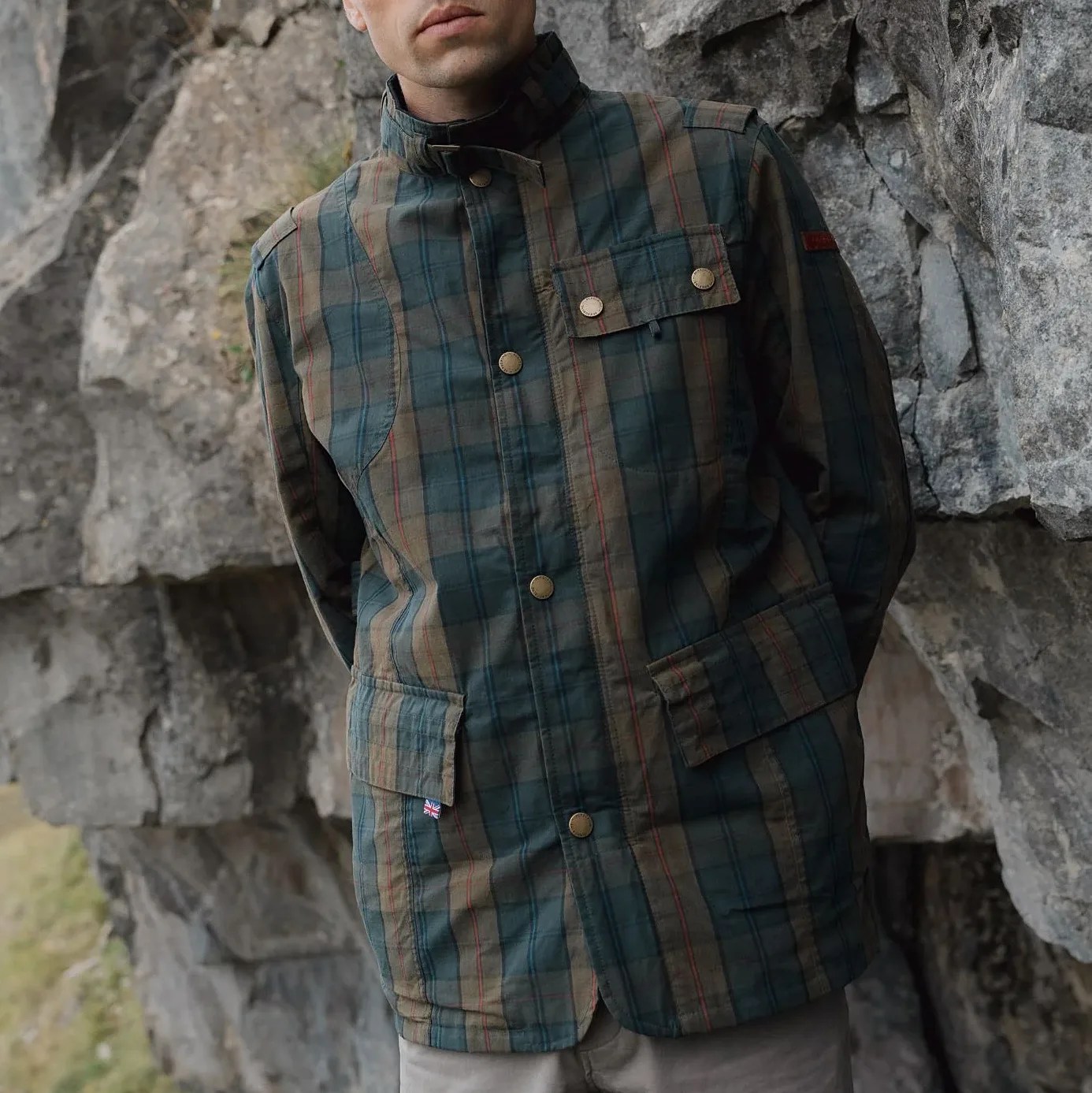 Green and brown plaid jacket with snap buttons and flap pockets worn by a person standing against a rocky background.