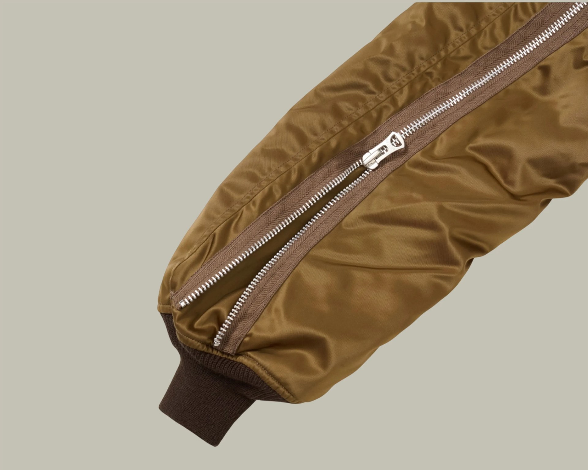 Brown jacket sleeve with a silver zipper and dark brown ribbed cuff on a beige background.