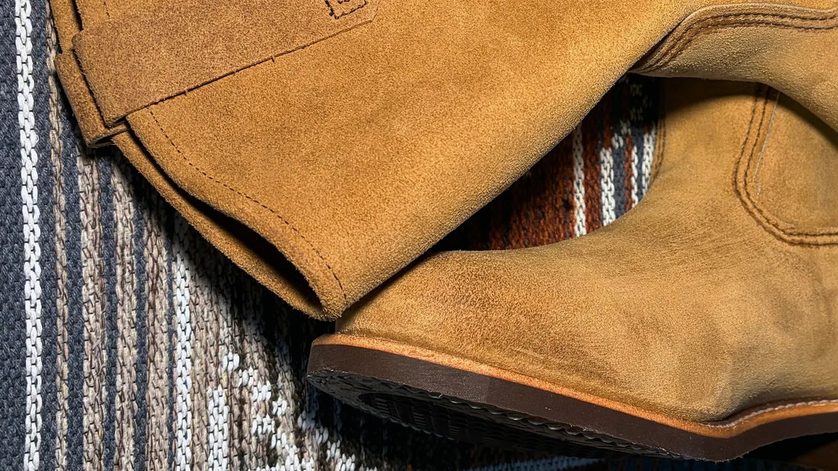 Close-up of tan suede boots with visible stitching on a striped woven fabric background.