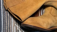 Close-up of tan suede boots with visible stitching on a striped woven fabric background.