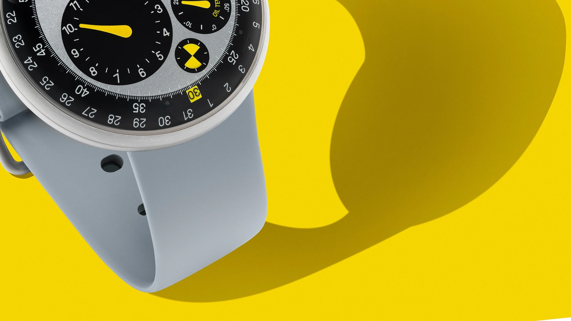 Close-up of a wristwatch with a silver case, black and gray dial, yellow hands, and a light gray silicone strap on a yellow background.