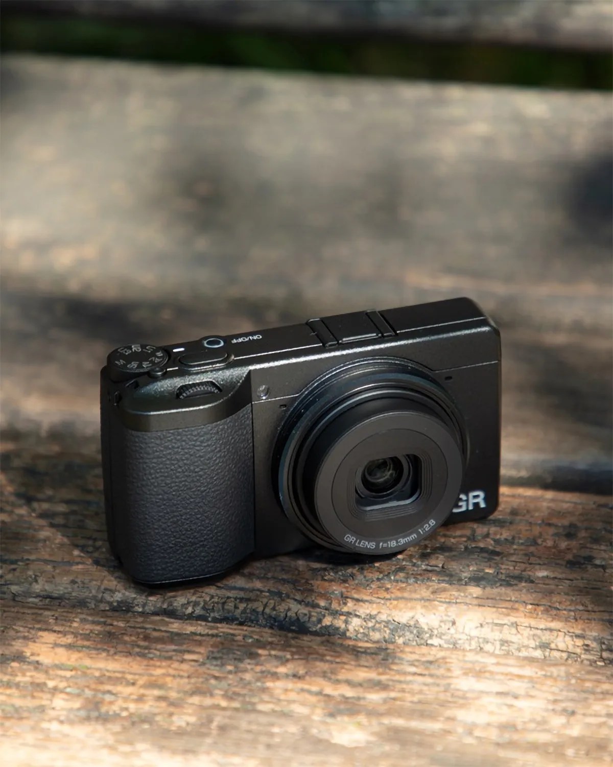 Compact black digital camera with textured grip and lens marked "GR LENS f=18.3mm 1:2.8" on a wooden surface.