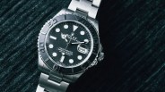 Silver Rolex Yacht-Master watch with black dial and date display on a textured dark surface.