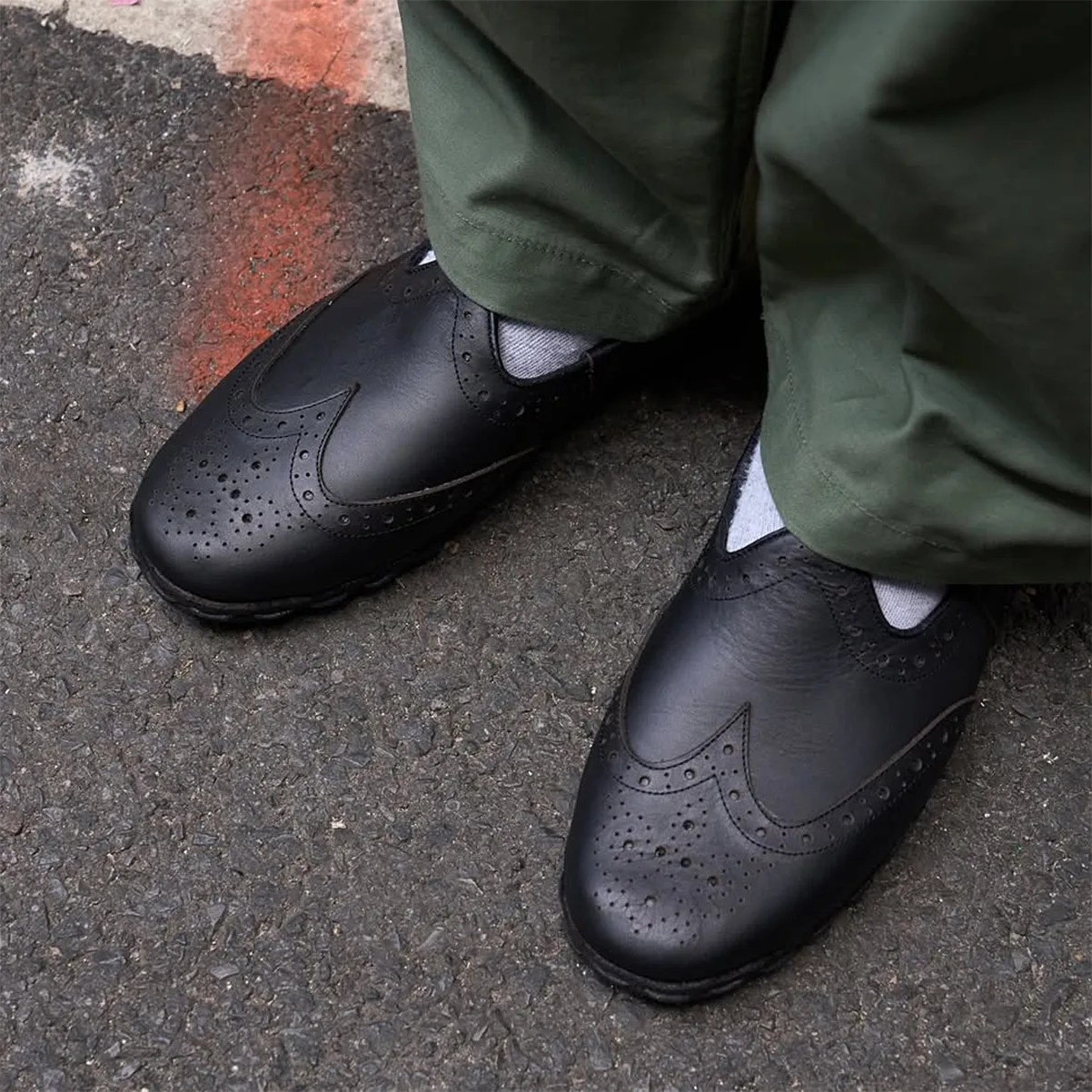 Black leather slip-on brogue shoes worn with light gray socks and green pants on asphalt ground.