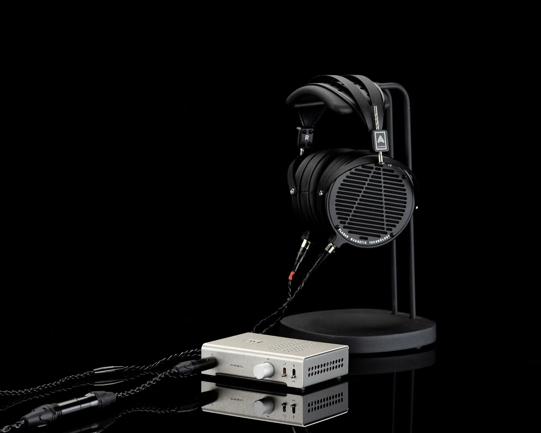 Black planar magnetic headphones on a stand connected to a silver audio amplifier with braided cables.