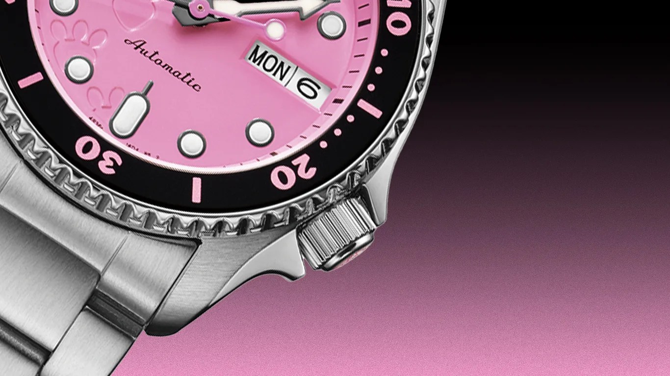 Close-up of a stainless steel watch with a pink dial, black bezel with pink markings, and a day-date display showing "MON 6".