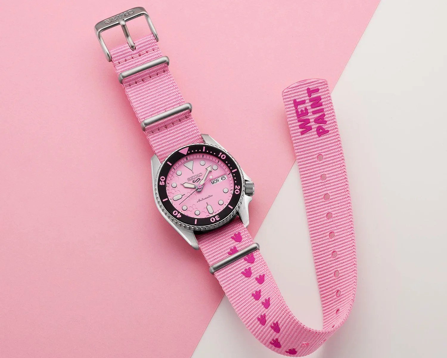 Seiko automatic watch with pink dial and black bezel on a pink fabric strap with "WET PAINT" text.
