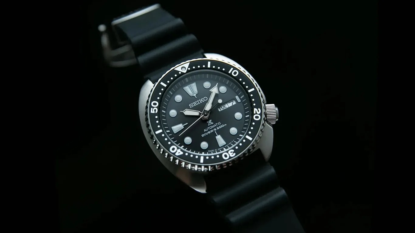 Black Seiko automatic diver's watch with round dial, luminous markers, and black rubber strap on a black background.