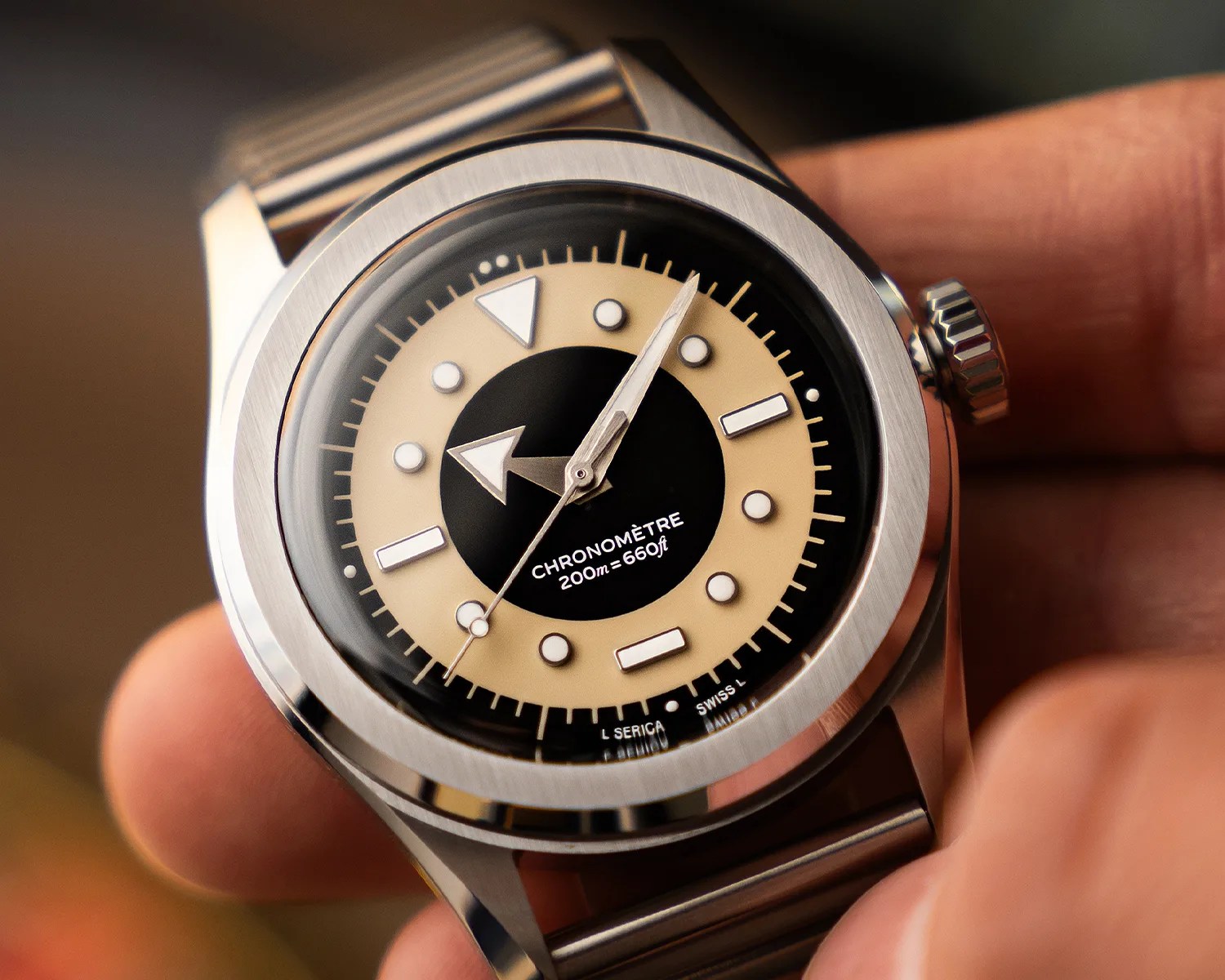 Close-up of a stainless steel wristwatch with a black and beige dial, luminous markers, and a metal bracelet held in a hand.