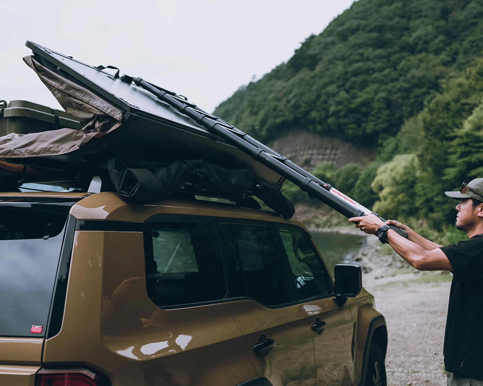 Man setting up a black rooftop tent on a brown SUV in a forested outdoor area.