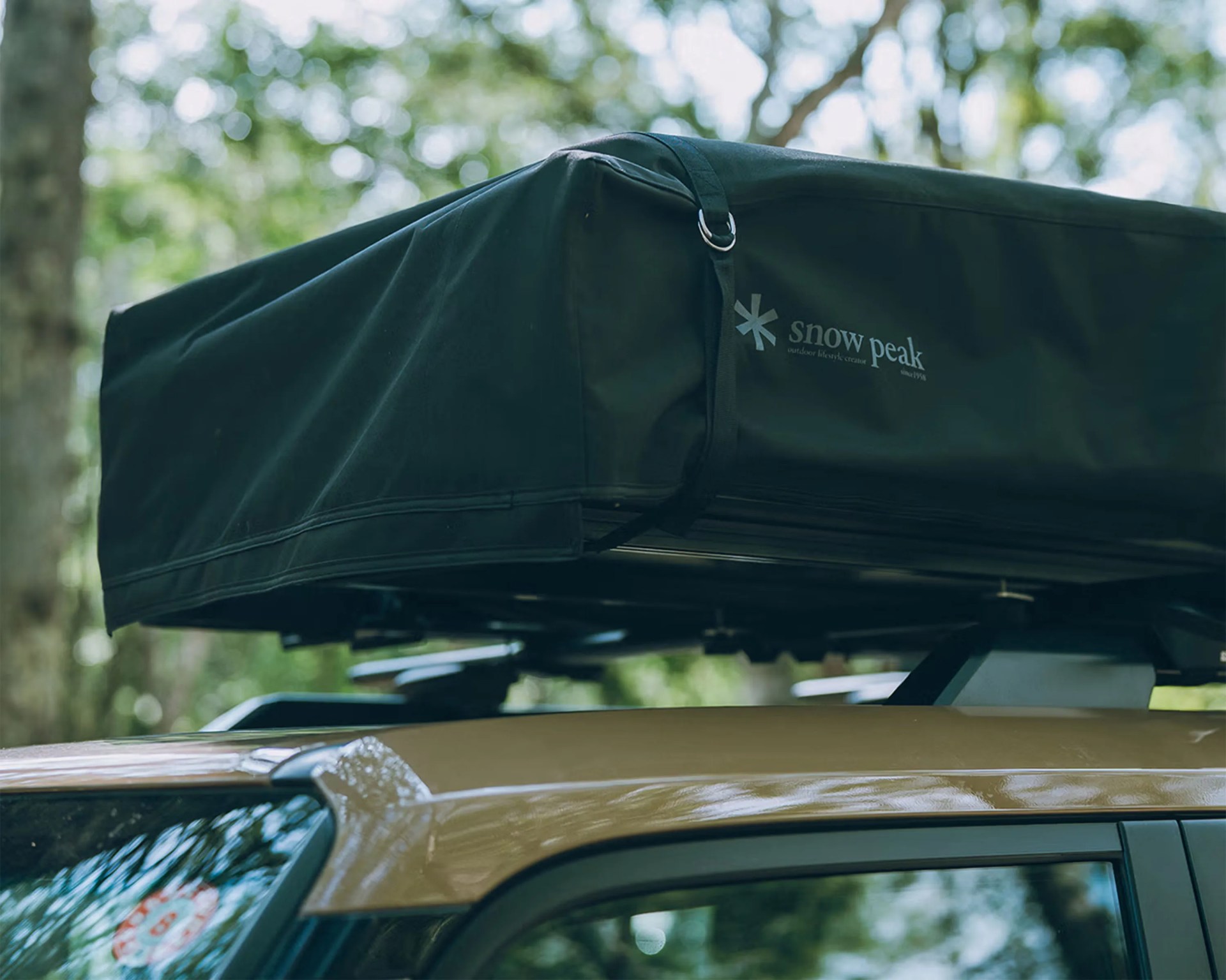 Black Snow Peak rooftop tent mounted on a tan vehicle roof rack in a forest setting.