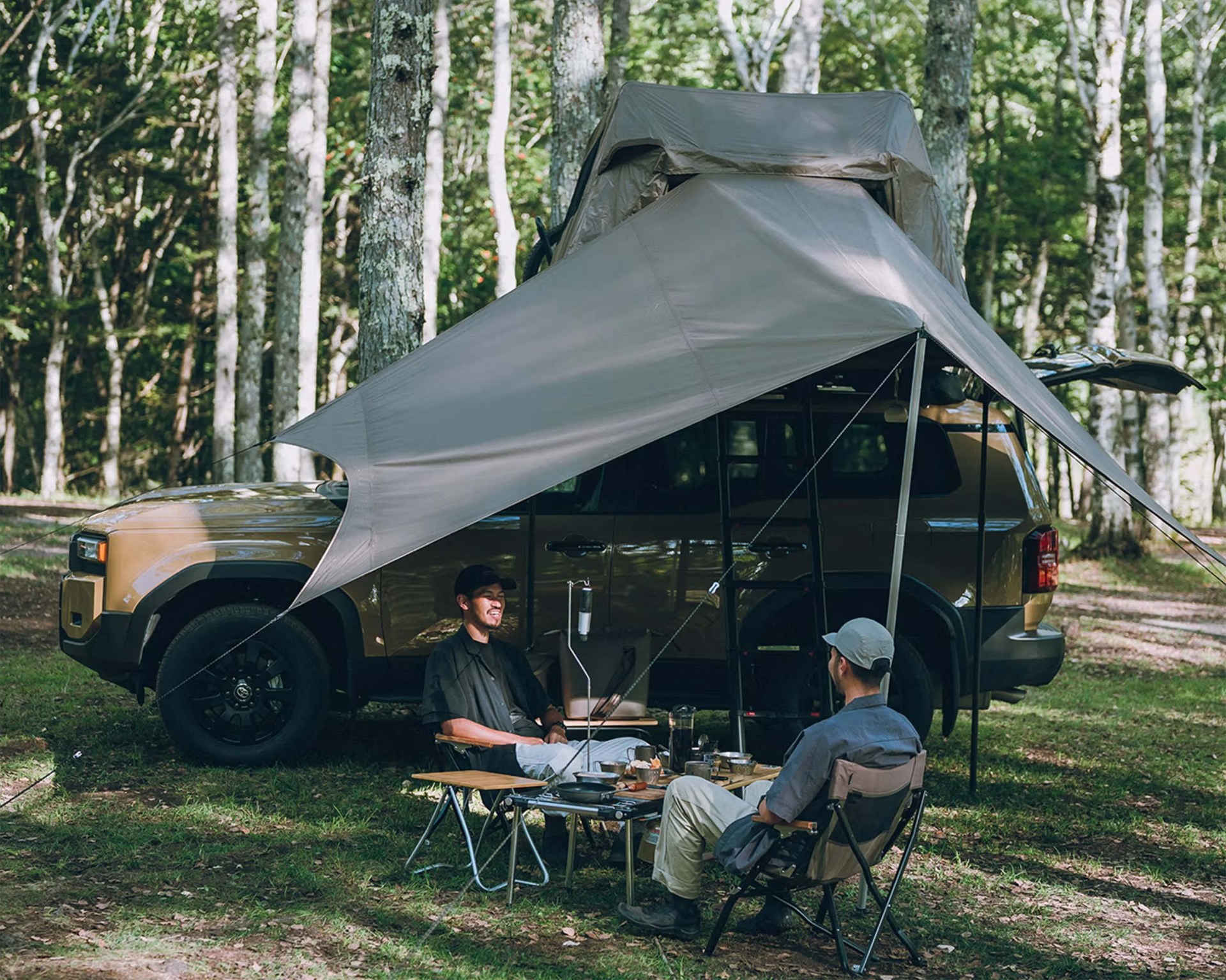 Brown SUV with rooftop tent and gray tarp awning set up in a forest with two people sitting at a camping table.