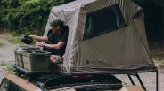 Man inside a taupe Snow Peak rooftop tent on a tan vehicle with green storage boxes on the roof rack.