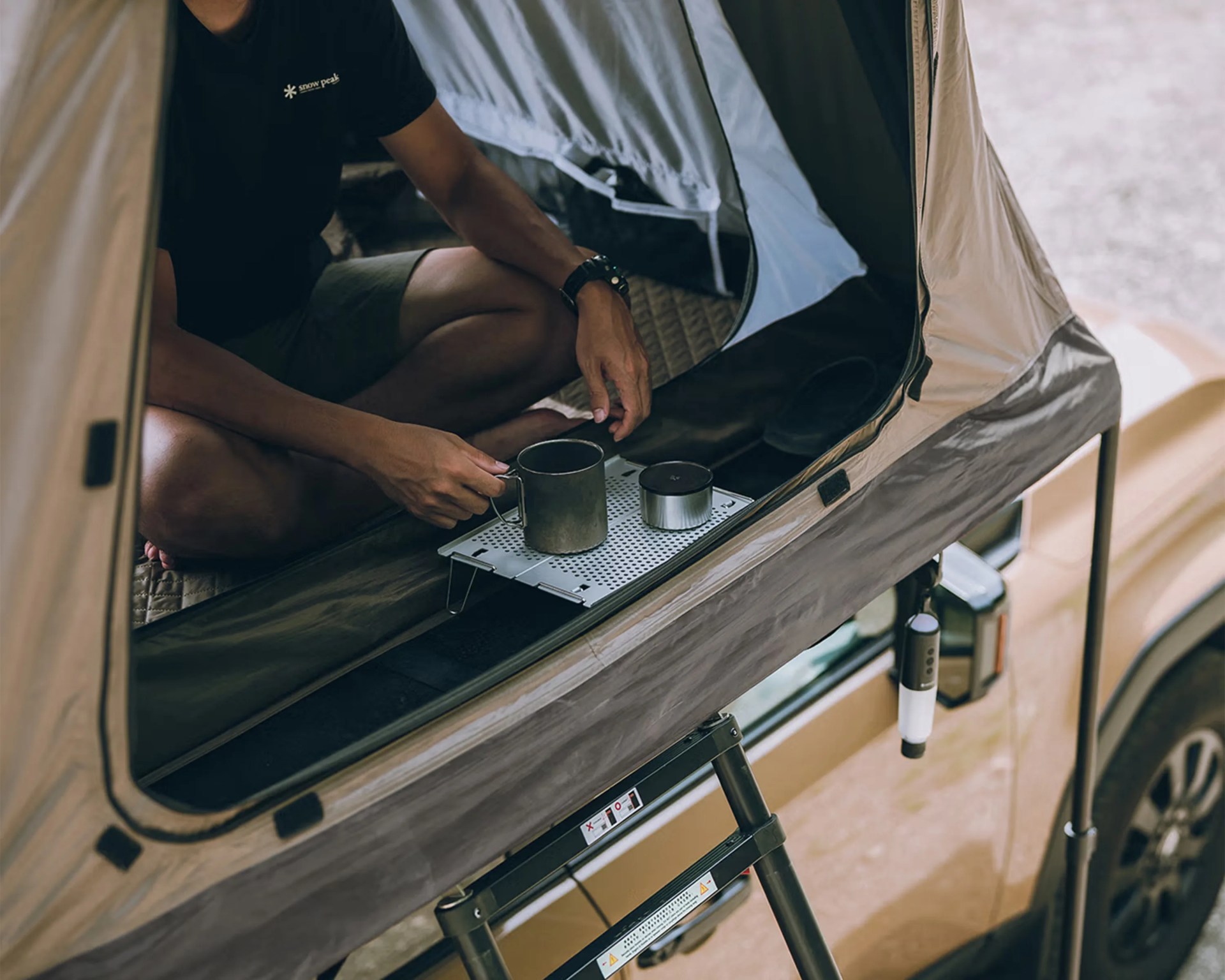 Person sitting inside a rooftop tent on a vehicle, holding a metal cup on a small foldable tray.
