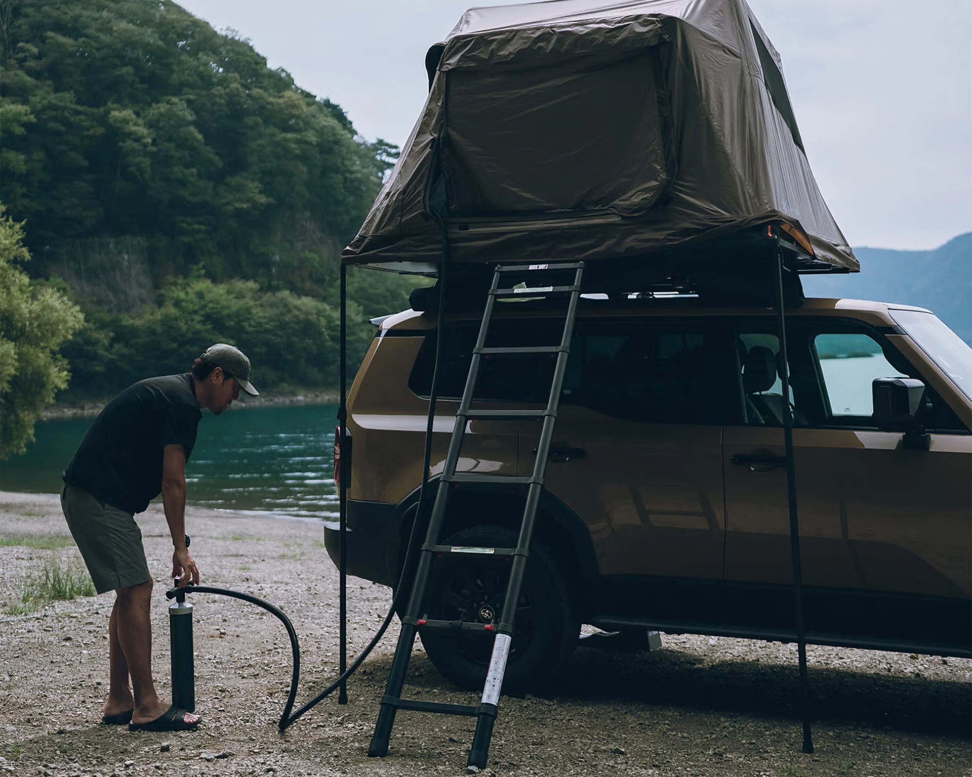 Man using a hand pump to inflate a rooftop tent mounted on a brown SUV near a lake.