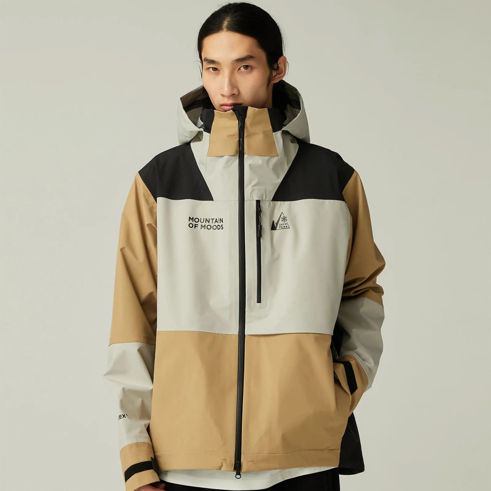 Beige, light gray, and black color-blocked hooded jacket with front zipper and chest pocket worn by a person.