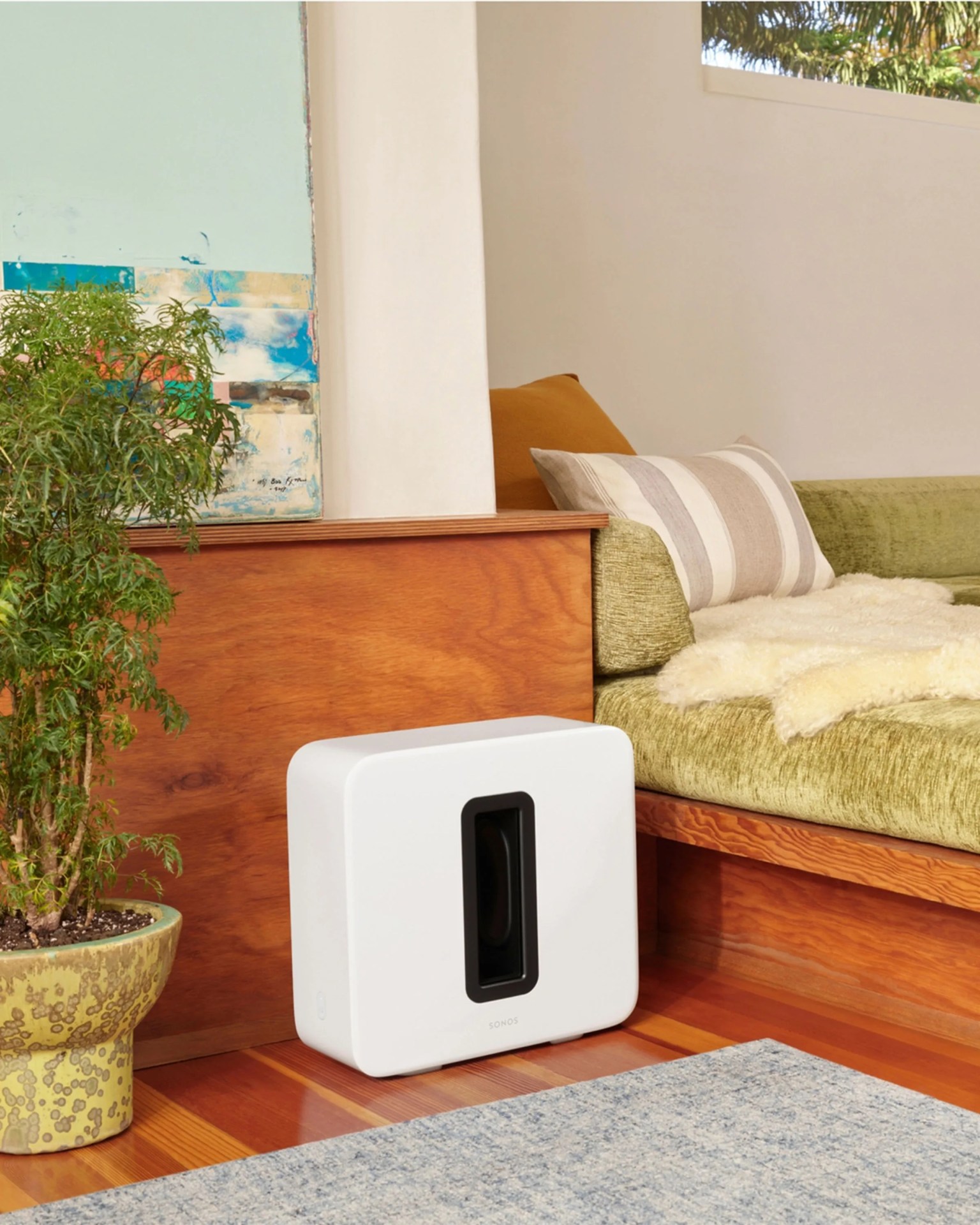 White Sonos subwoofer speaker on wooden floor next to green couch with pillows and a potted plant.