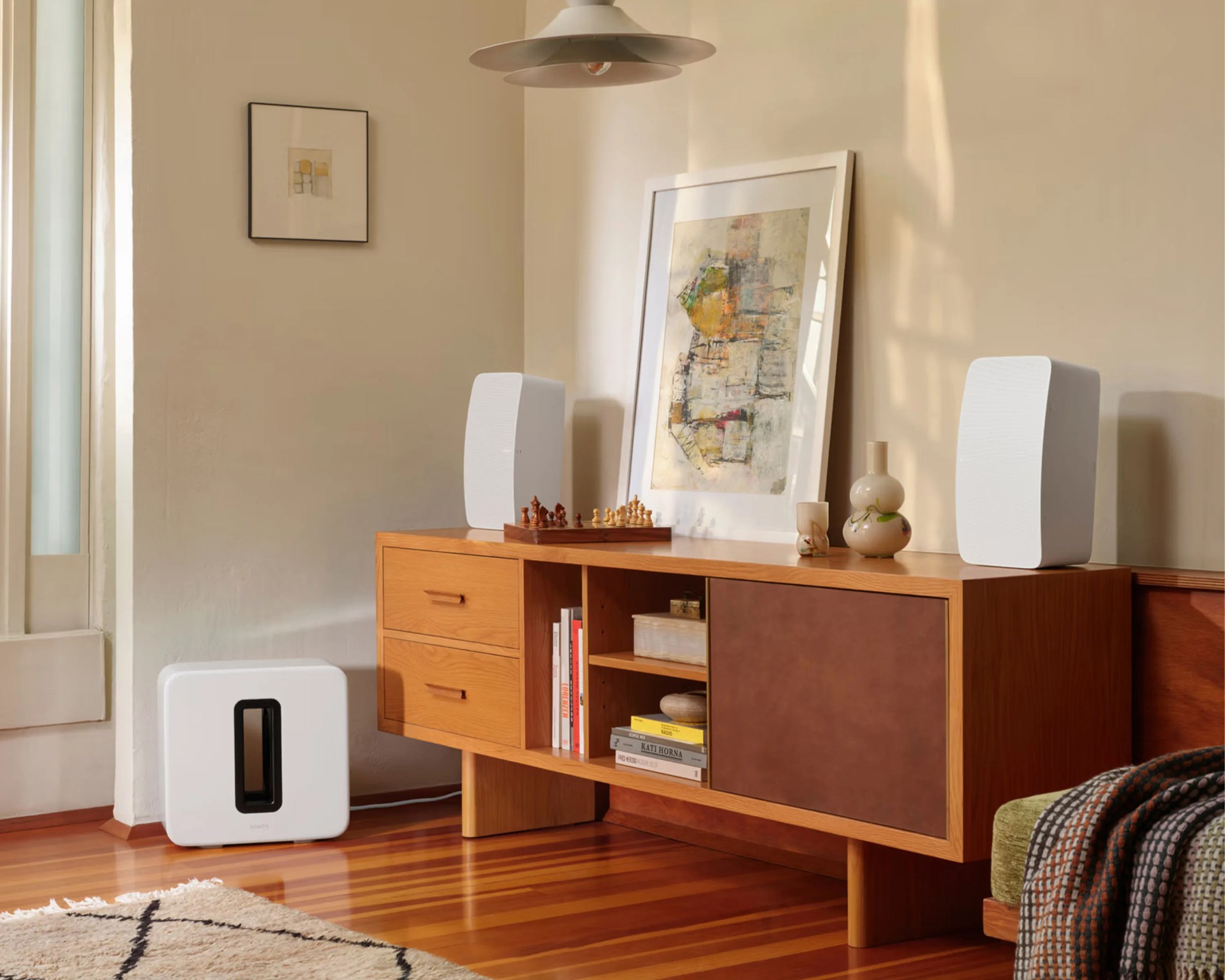 Wooden sideboard with white bookshelf speakers, chess set, framed artwork, and ceramic vases in a living room.