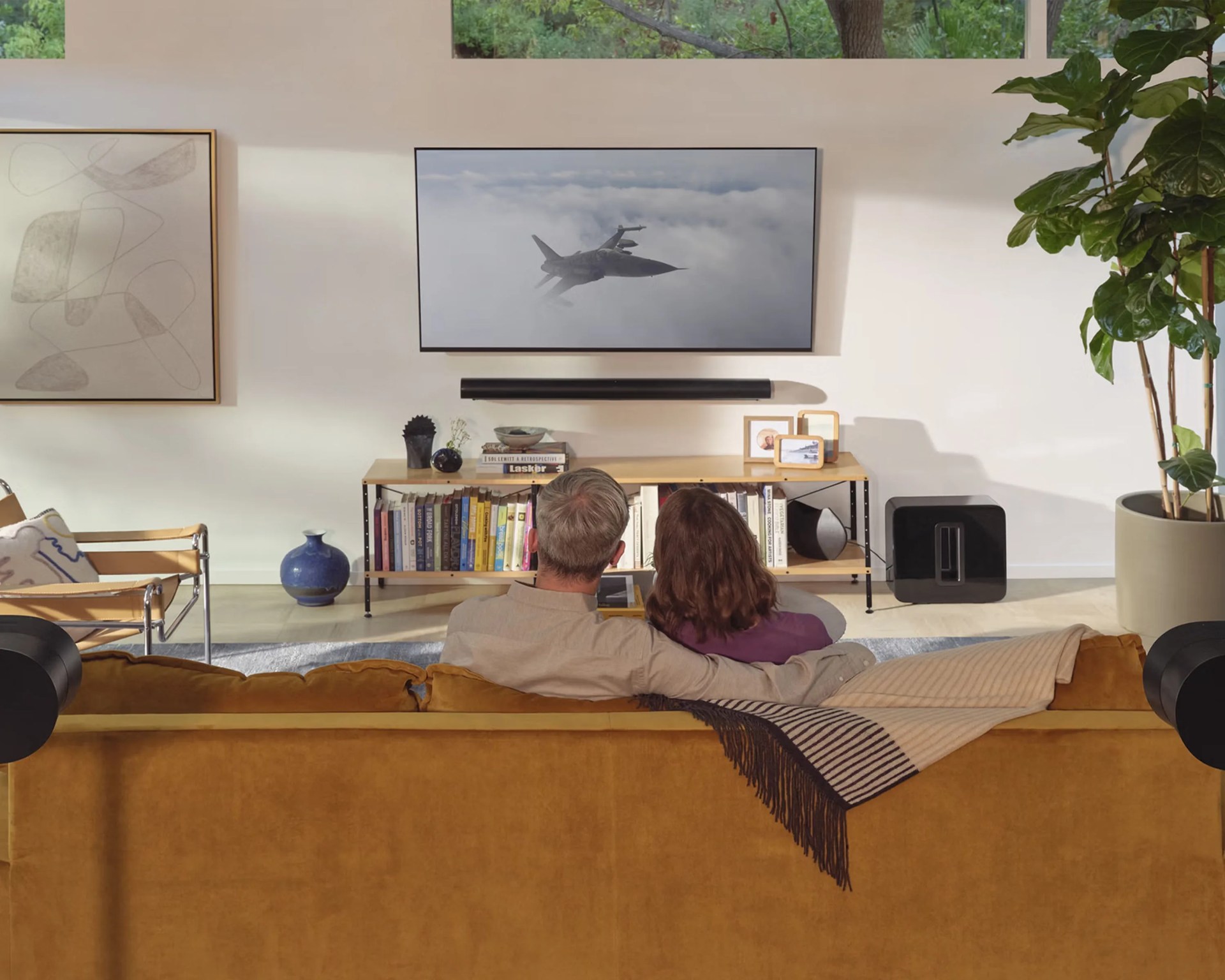 Couple sitting on a mustard yellow sofa watching a jet fighter on a wall-mounted TV in a modern living room.