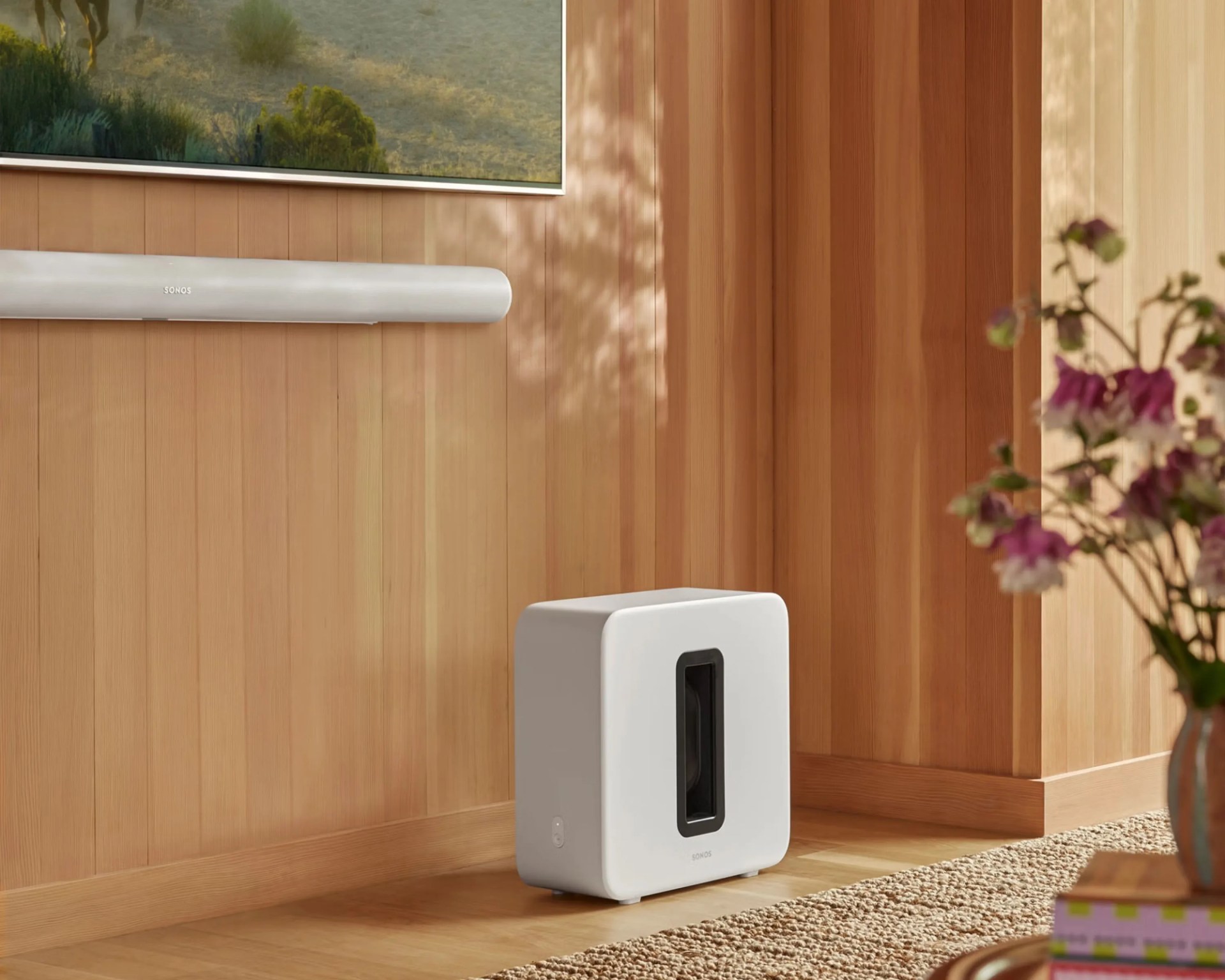 White Sonos subwoofer speaker on a wooden floor next to a beige rug and a wall-mounted Sonos soundbar.