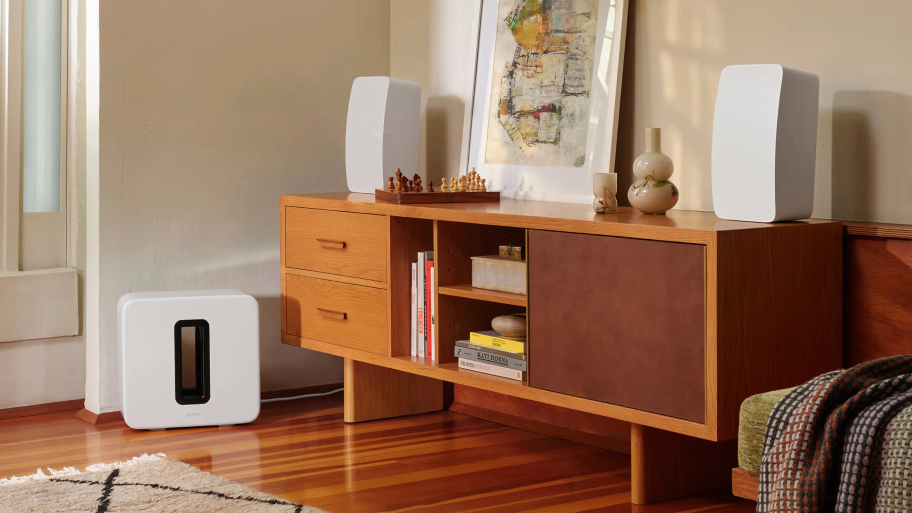 White Sonos subwoofer speaker on wooden floor next to wooden sideboard with white bookshelf speakers and decorative items.