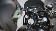 Close-up of a black motorcycle dashboard with a circular digital display showing music controls and a gloved hand pressing a button.
