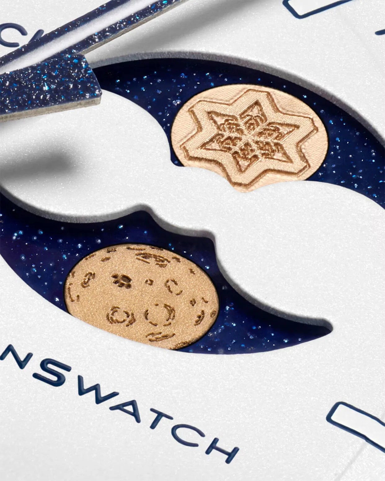 Close-up of a white watch face with blue glittery hands and two gold decorative elements, one star-shaped and one circular, on a dark blue background.
