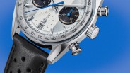Close-up of a silver chronograph watch with black subdials, blue hands, tachymeter scale, and black perforated leather strap.