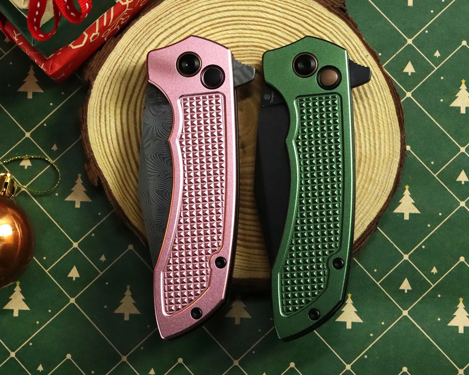 Two folding knives with textured handles, one pink and one green, placed on a wooden slice with a green patterned background.