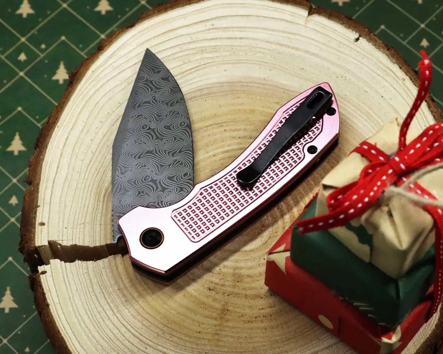 Folding knife with a patterned blade and pink textured handle on a wooden tree slice next to small wrapped gifts.
