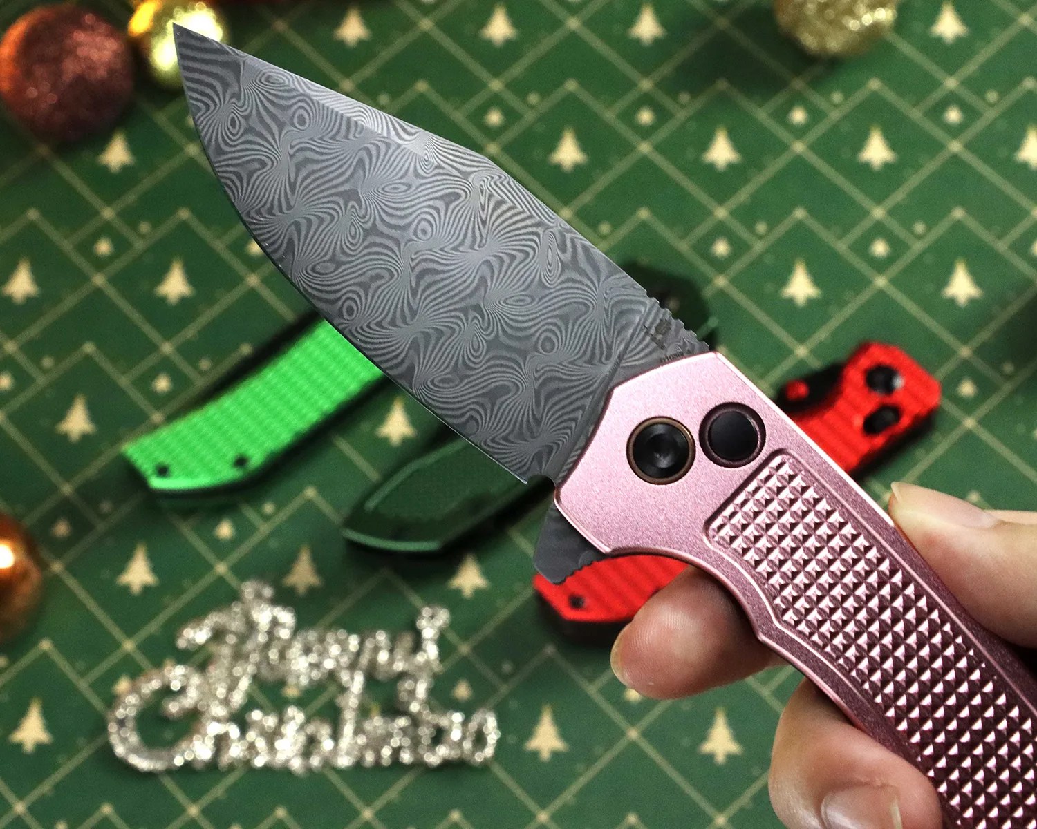 Hand holding a folding knife with a patterned Damascus steel blade and textured pink handle over a green Christmas-themed background.