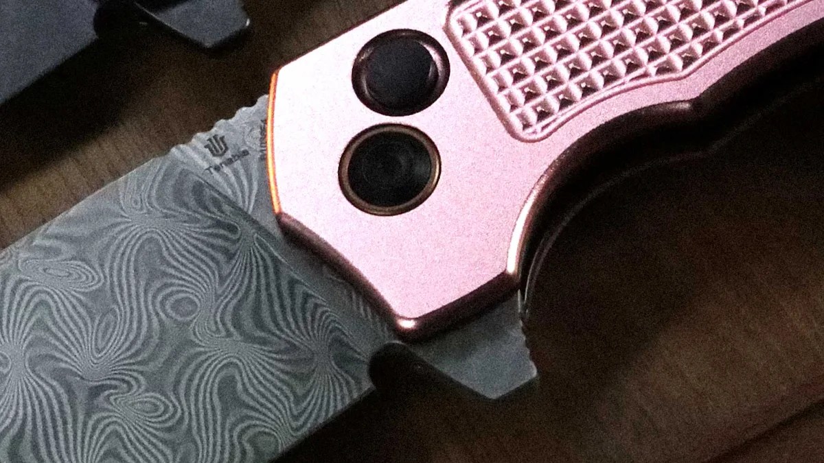 Close-up of a folding knife with a patterned Damascus steel blade and a rose gold textured handle.